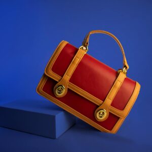 Stylish red and tan leather handbag with handle and gold accents on vibrant blue backdrop.