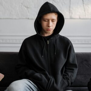 Teenager in a black hoodie sitting on a couch, reflecting on thoughts indoors.