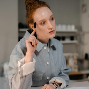 A redheaded fashion designer in a gray shirt thinking while holding a pen at a wooden table with papers.