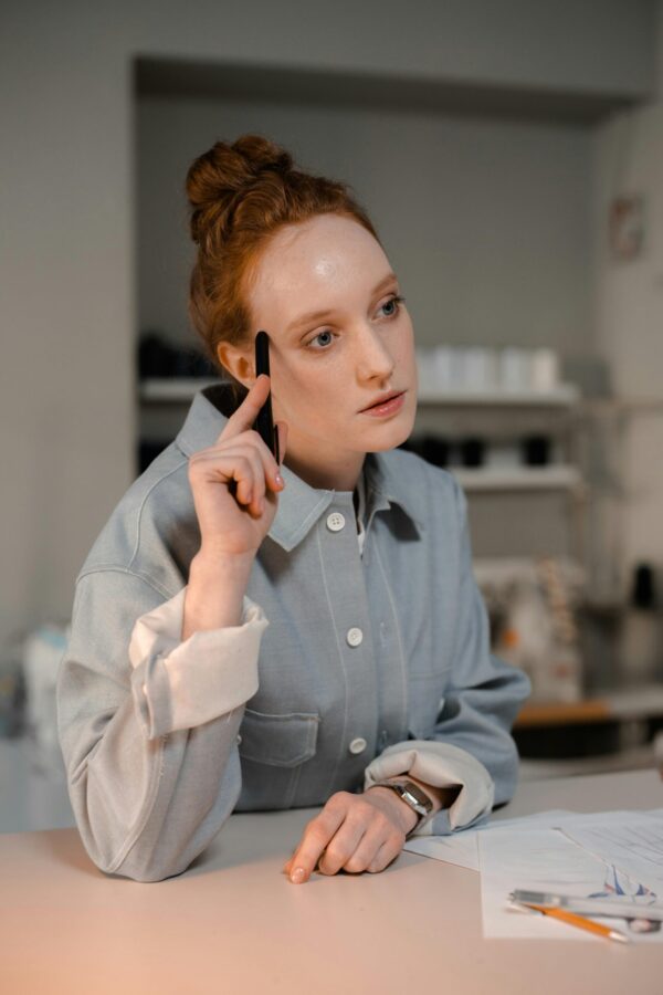 A redheaded fashion designer in a gray shirt thinking while holding a pen at a wooden table with papers.